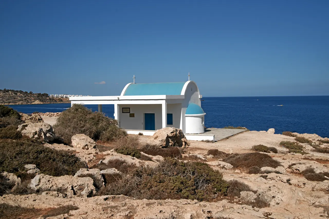 White chapel with a blue dome and roof on a rocky headland, low scrub in front and the open sea behind