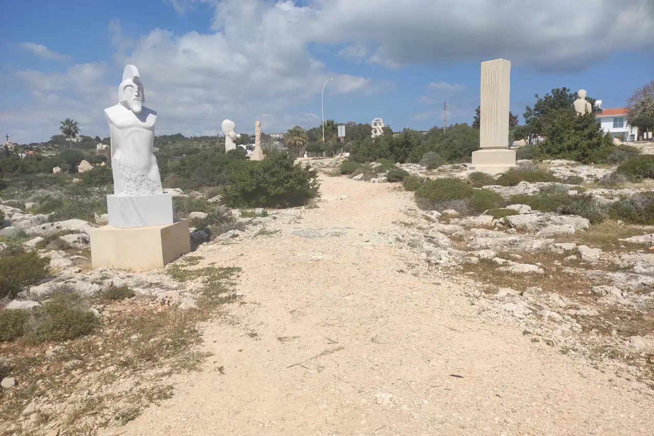 A white marble bust beside a sandy path through low shrubs, with more sculptures in the distance