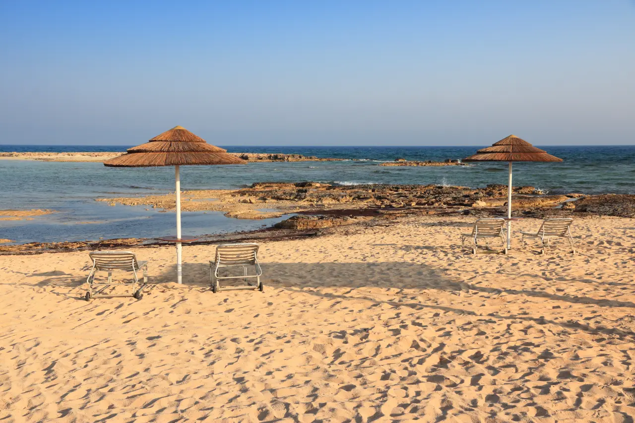 Sandy beach with two thatched sunshades and loungers, rocks and shallow sea behind