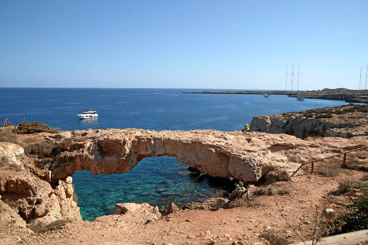 Natural stone arch over turquoise water on the rocky coast, a motor yacht on the open sea behind it