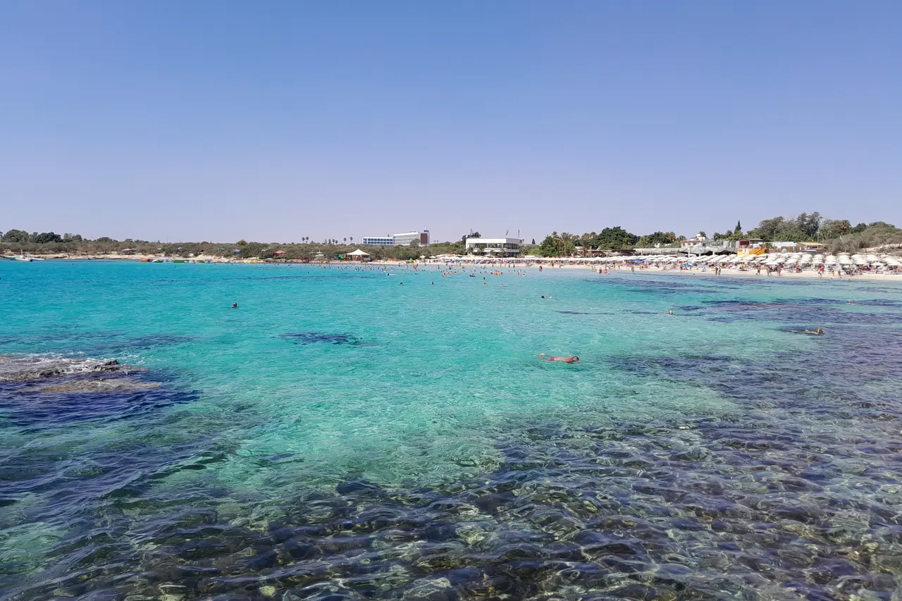Clear turquoise shallows in a bay, with the beach and sunshades in the distance
