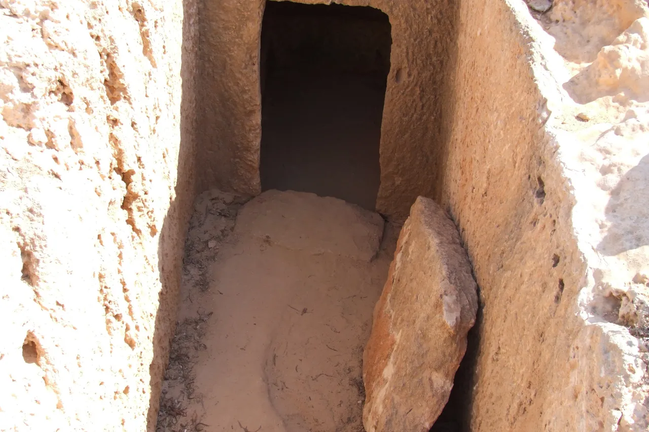 Entrance of a tomb cut into pale limestone, with a stone slab leaning in the opening