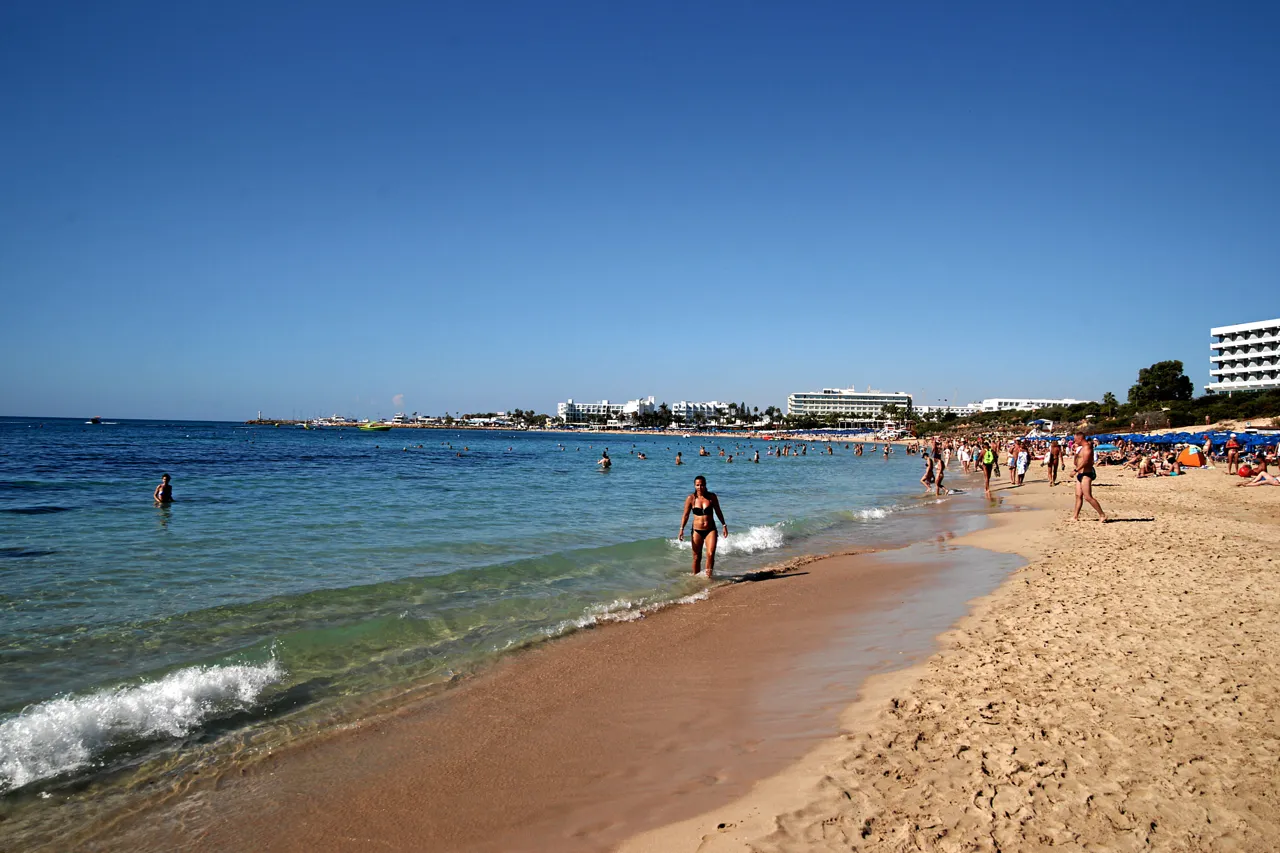 Long sandy beach with bathers in clear, shallow water and hotels along the shore in the distance