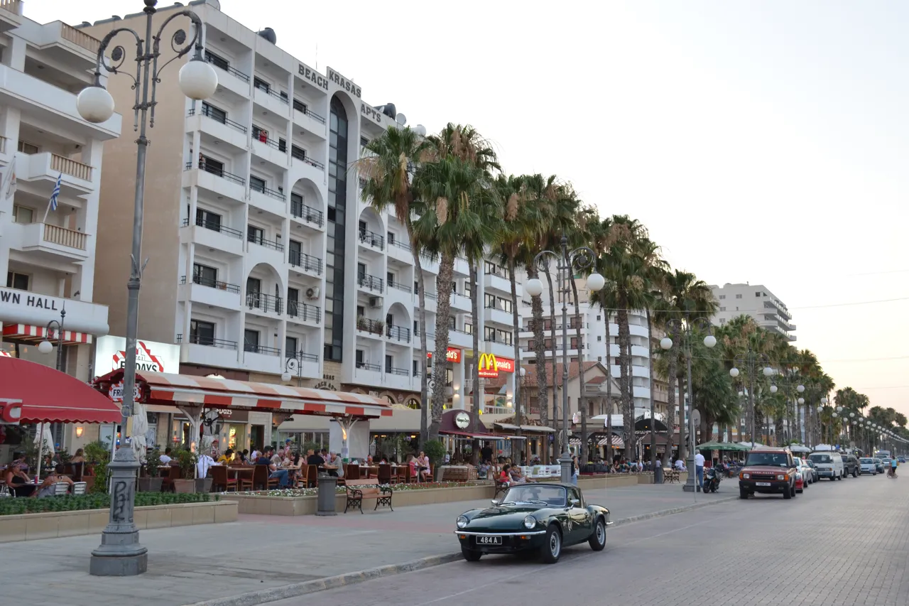 Palm-lined seafront street with café terraces and hotels on the left and a vintage car in the foreground