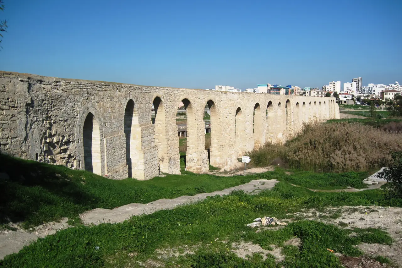 Long stone aqueduct with many arches over grass, a blue sky and white buildings in the distance