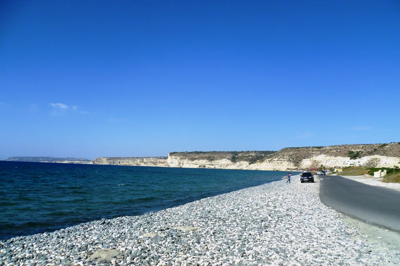 Pebble beach beside a road, with dark blue sea and pale cliffs along the coast under a clear sky