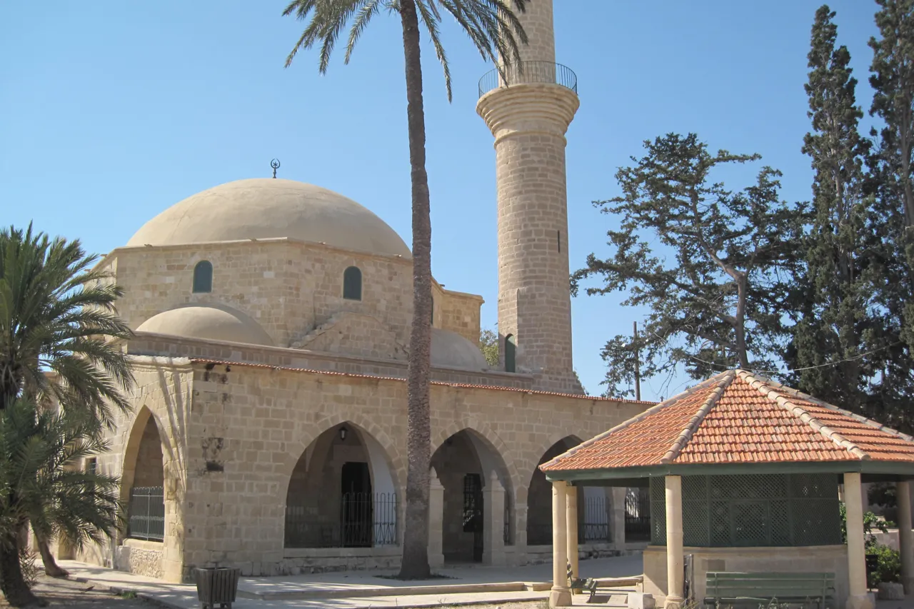 Stone mosque with a dome and a tall minaret, palms and pines, and a tiled shelter in the foreground