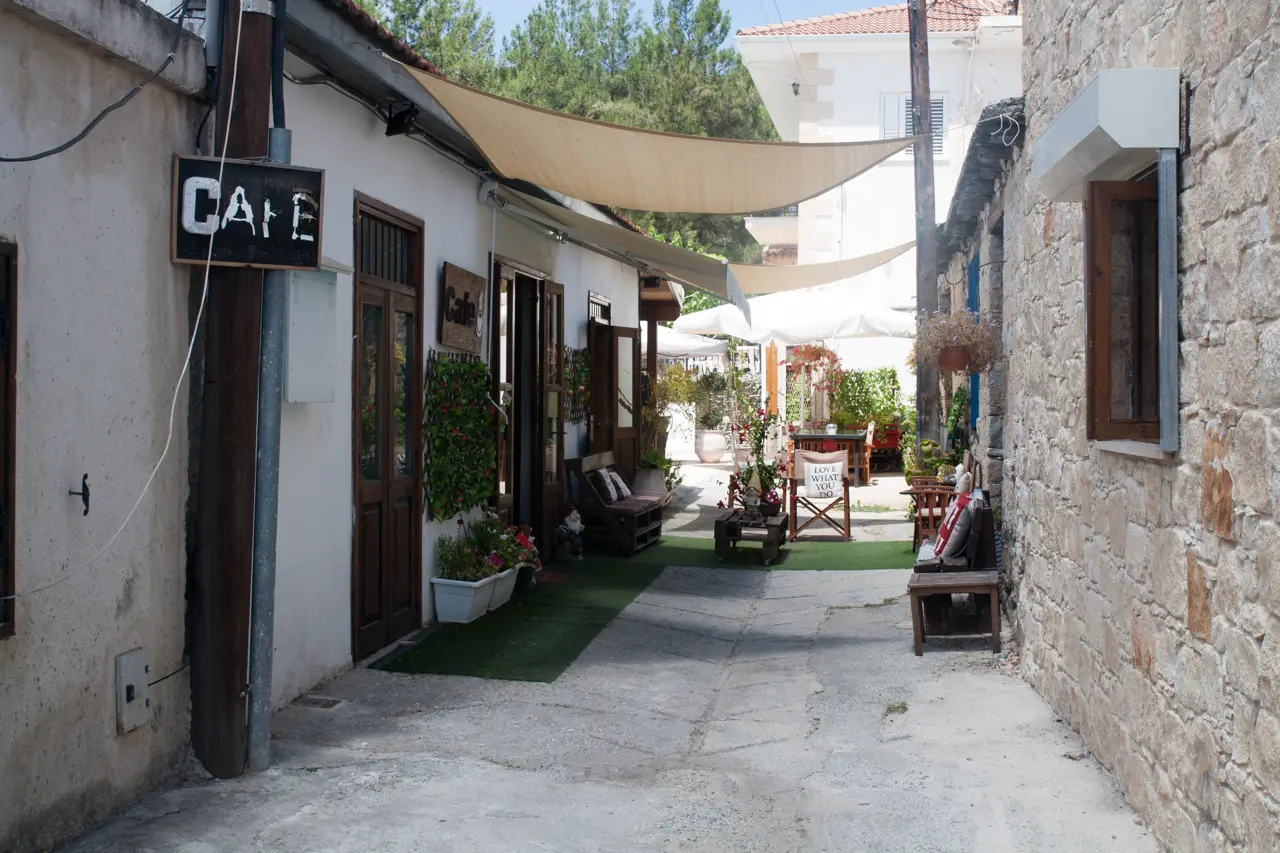 Quiet lane between old houses, with a café sign, shade sails and tables in a courtyard