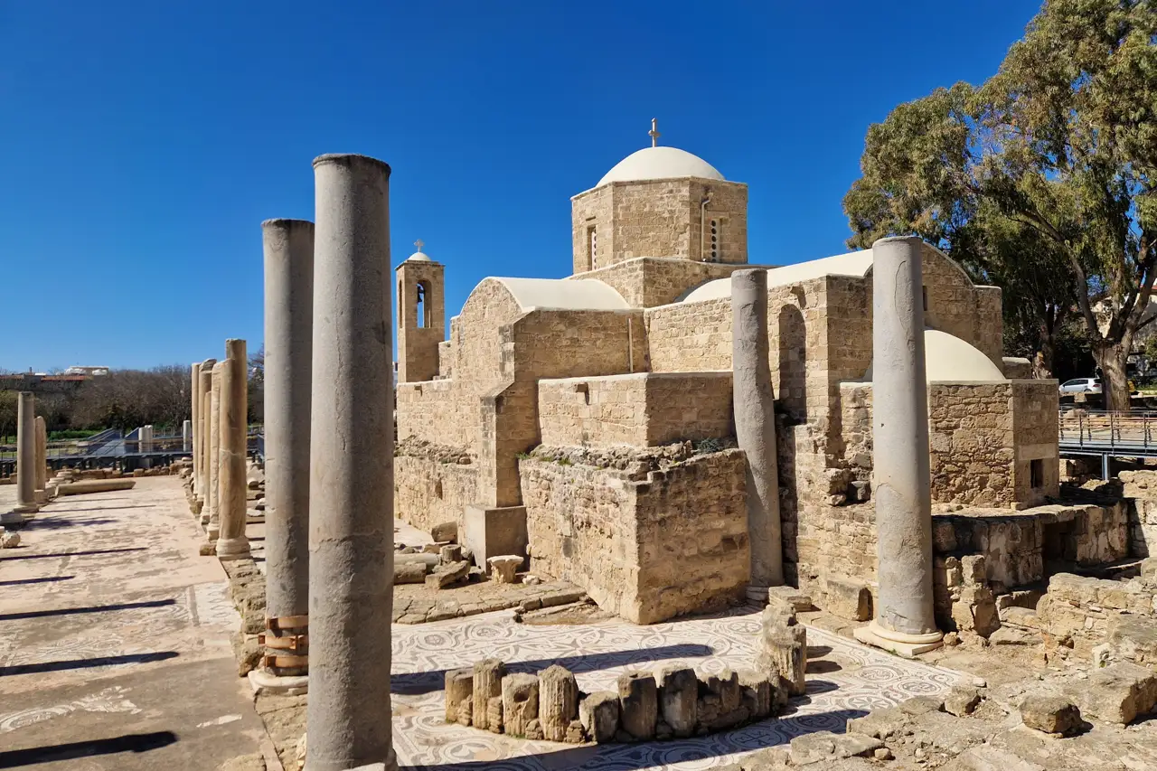 Small domed stone church behind rows of tall columns and low basilica ruins, in bright sunshine