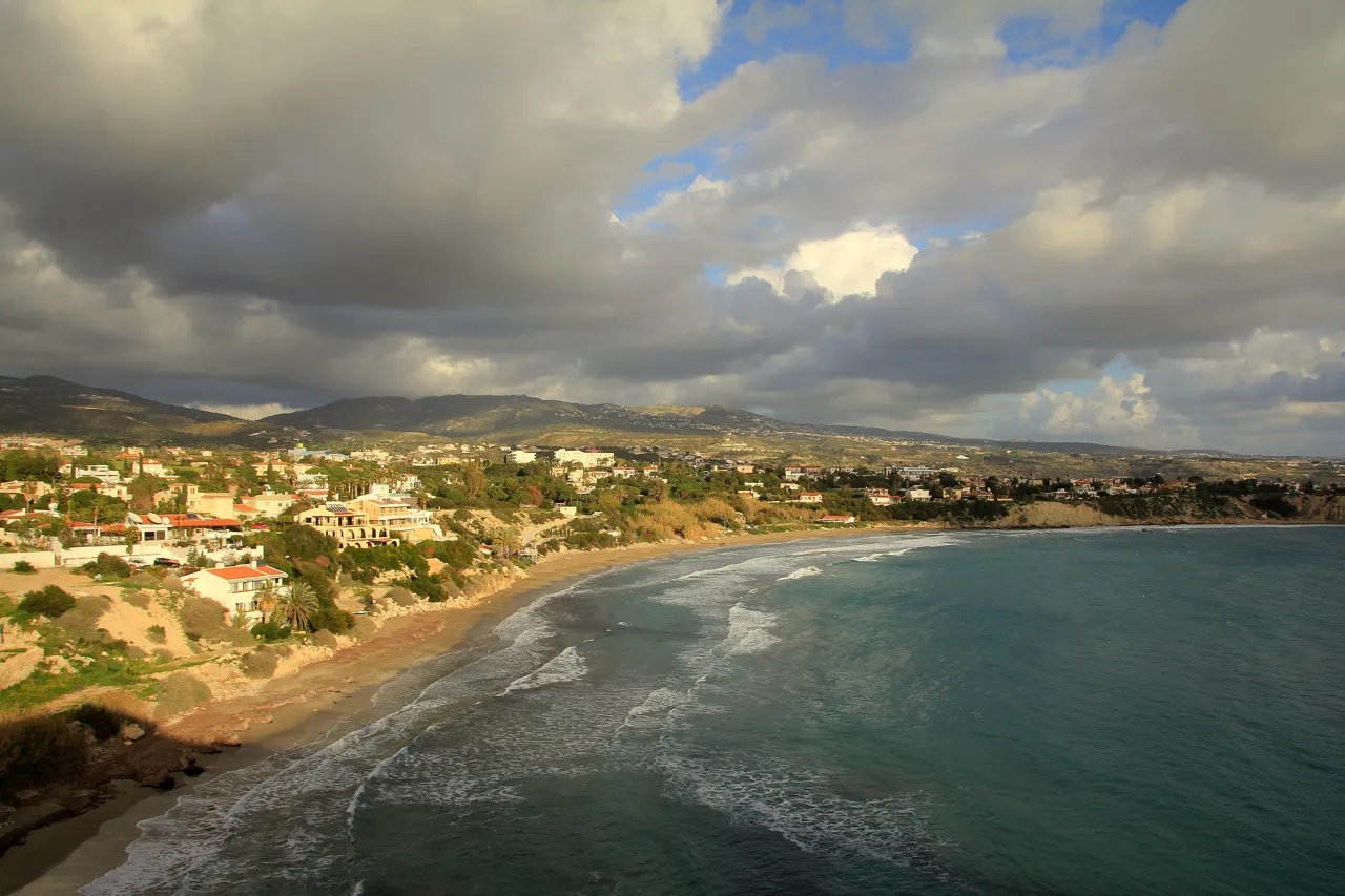 Curved sandy bay seen from above, with waves breaking, houses along the shore and dark clouds over the hills