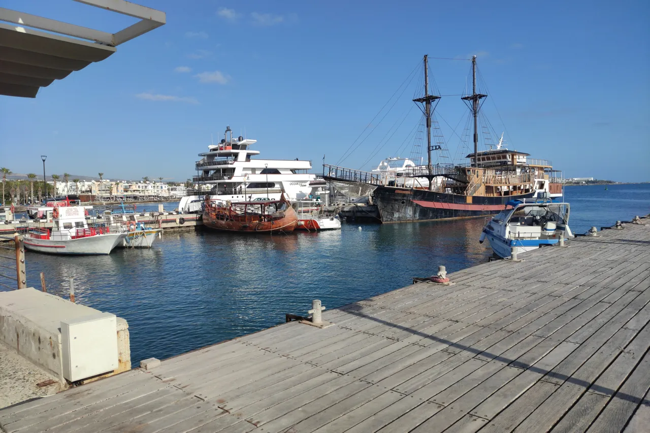 Wooden quay at Paphos harbour with moored boats, a tall-masted ship and a white tour boat under a blue sky