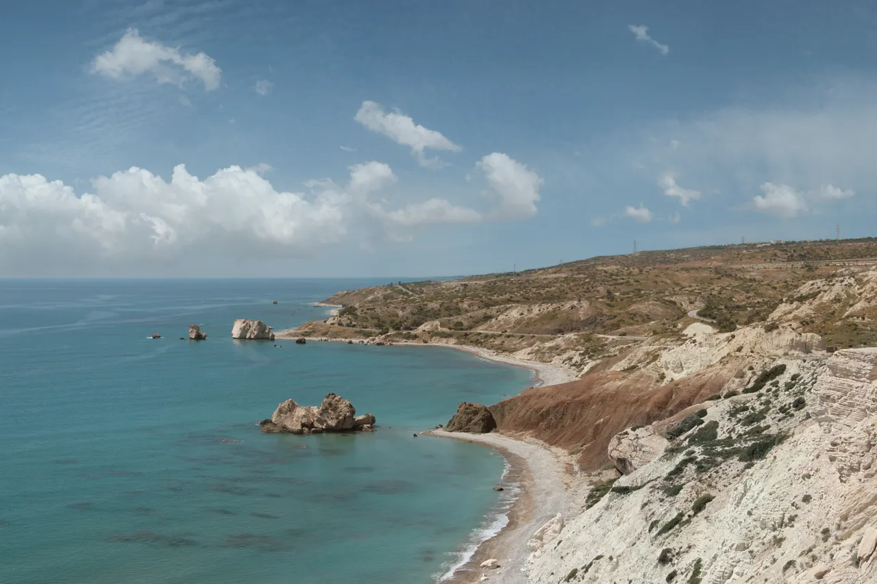 Turquoise bay with two rocks in the water, pale cliffs and a thin sandy shore under a partly clouded sky