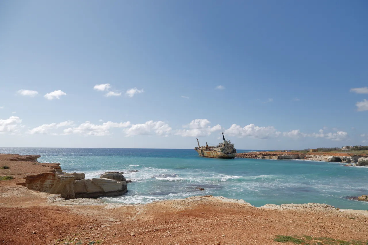 Rocky red-brown coast above a turquoise bay with surf, and a rusty wreck aground near the shore under a blue sky