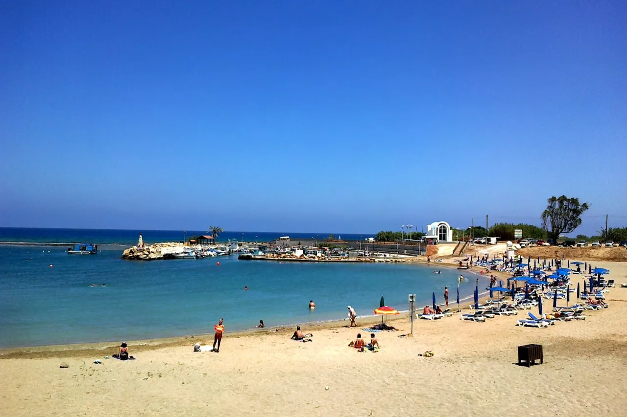Wide sandy beach with calm water, a small harbour with boats in the middle distance and a white chapel beside sunbeds