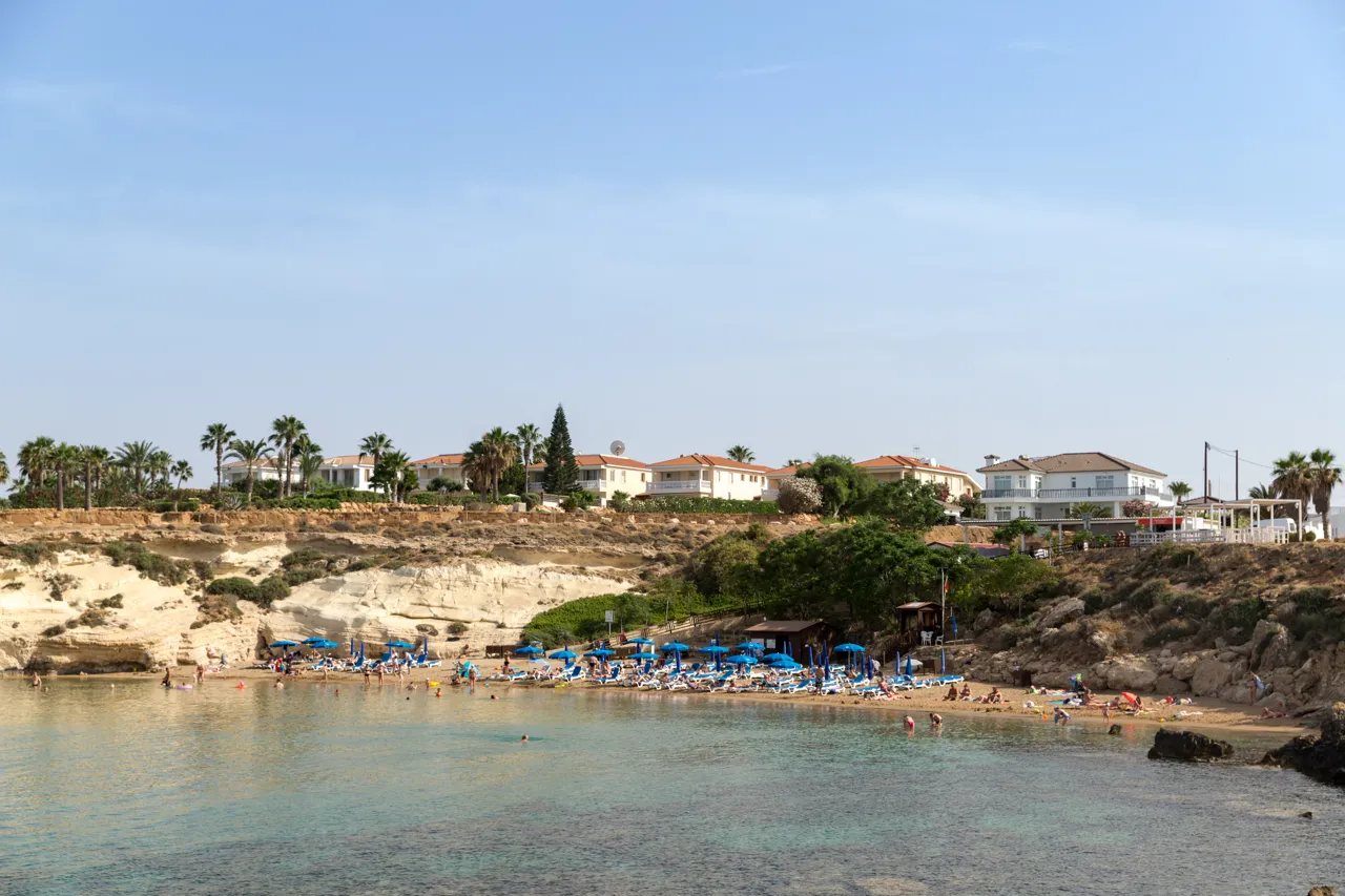 Small bay with a narrow sandy beach, blue umbrellas and sunbeds below white limestone cliffs and holiday houses