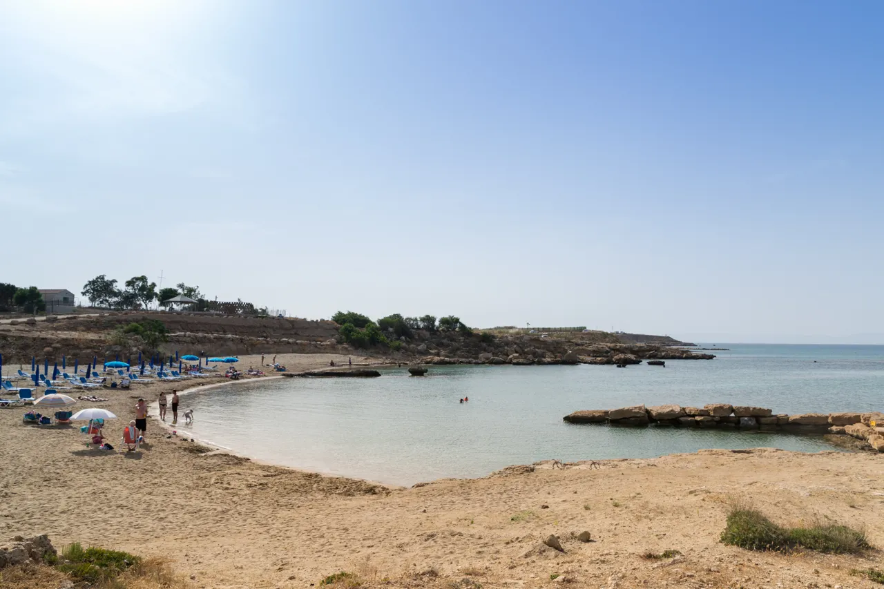 Shallow sandy bay with calm water, a few sunbeds on the left, low rocks on the right and a few bathers
