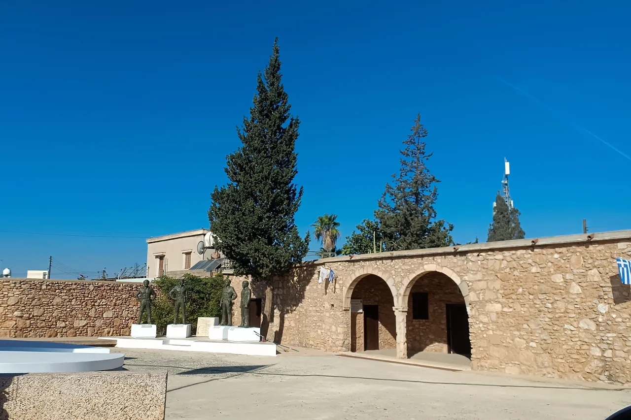 Stone building with two arched doorways beside a plaza with bronze figures on white plinths and a tall cypress