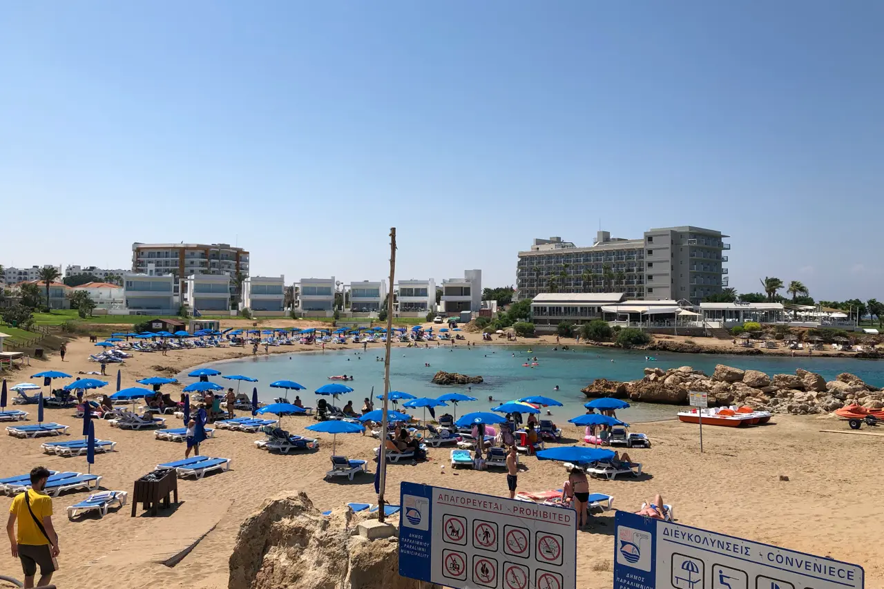 Small sandy bay with rows of blue umbrellas and sunbeds, rocks on the right and hotels behind, under a clear sky