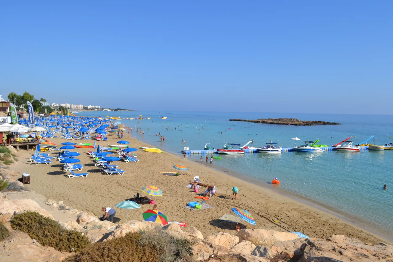 Sandy beach with rows of blue sunbeds, swimmers, moored boats and a low rocky island offshore