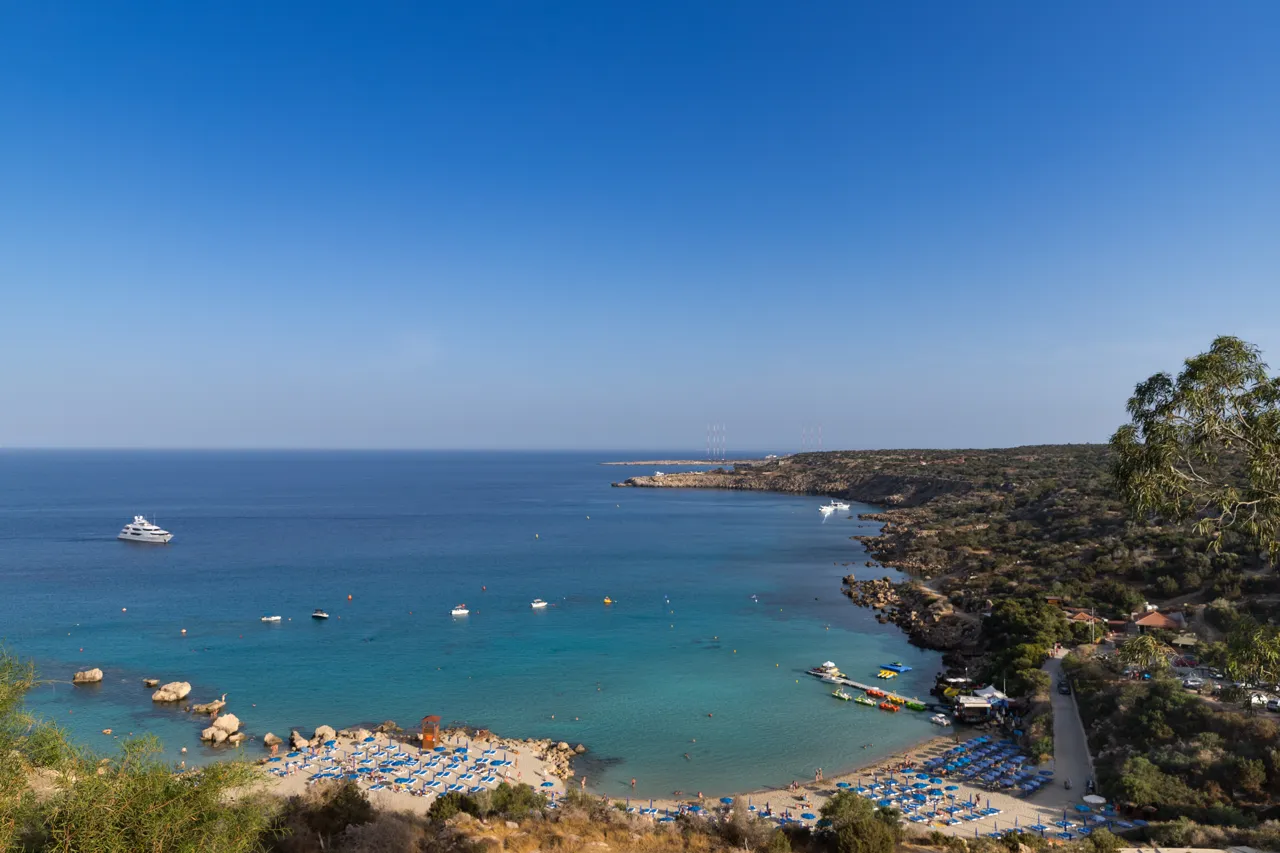 Hillside view over a turquoise bay with blue sunbeds on the sand, a white yacht and a wooded headland
