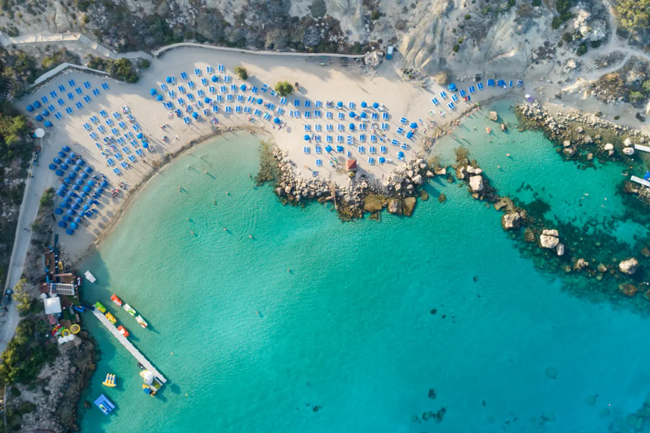 Aerial view of Konnos Bay: a sandy beach with rows of blue sunbeds, a wooden jetty and turquoise water