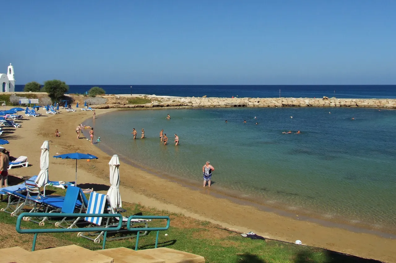 Louma Beach: a shallow sandy bay with swimmers, sunbeds on a grassy bank and a small white chapel at left