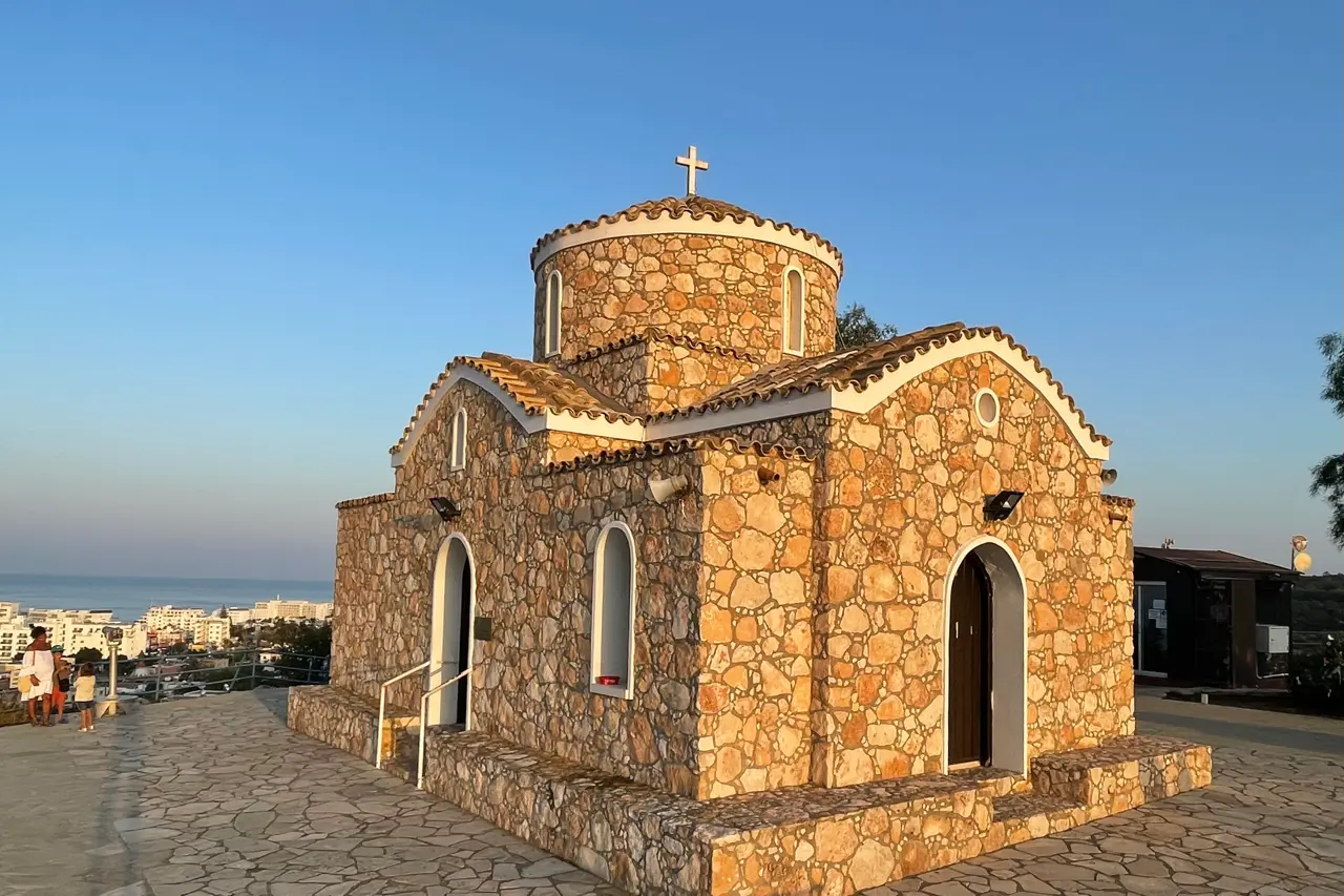 Small stone chapel with a domed roof and arched doors on a paved terrace, with the town and the sea behind
