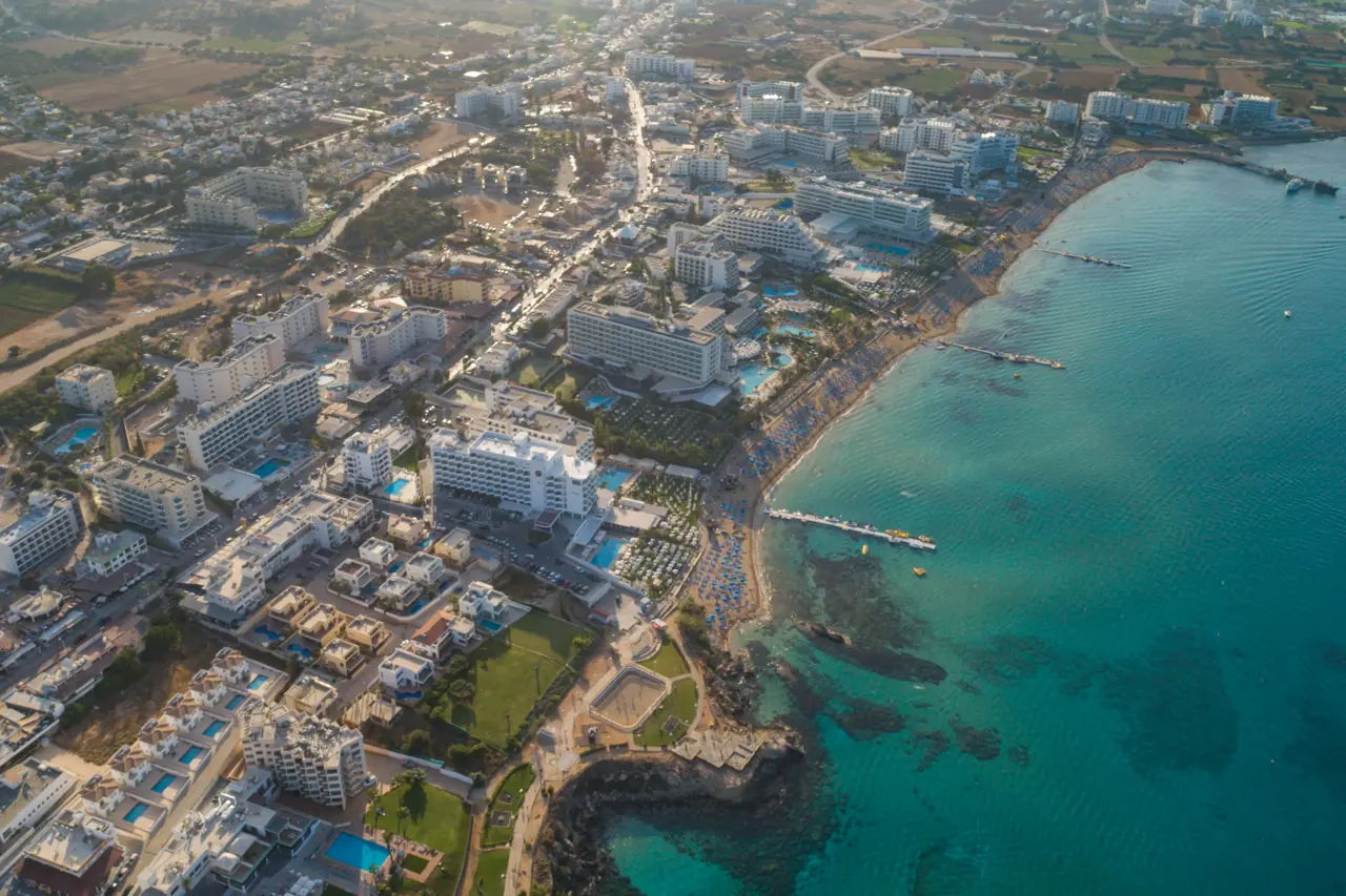 Aerial view along the Vrysi strip, with jetties, hotels and streets behind the beach and turquoise water