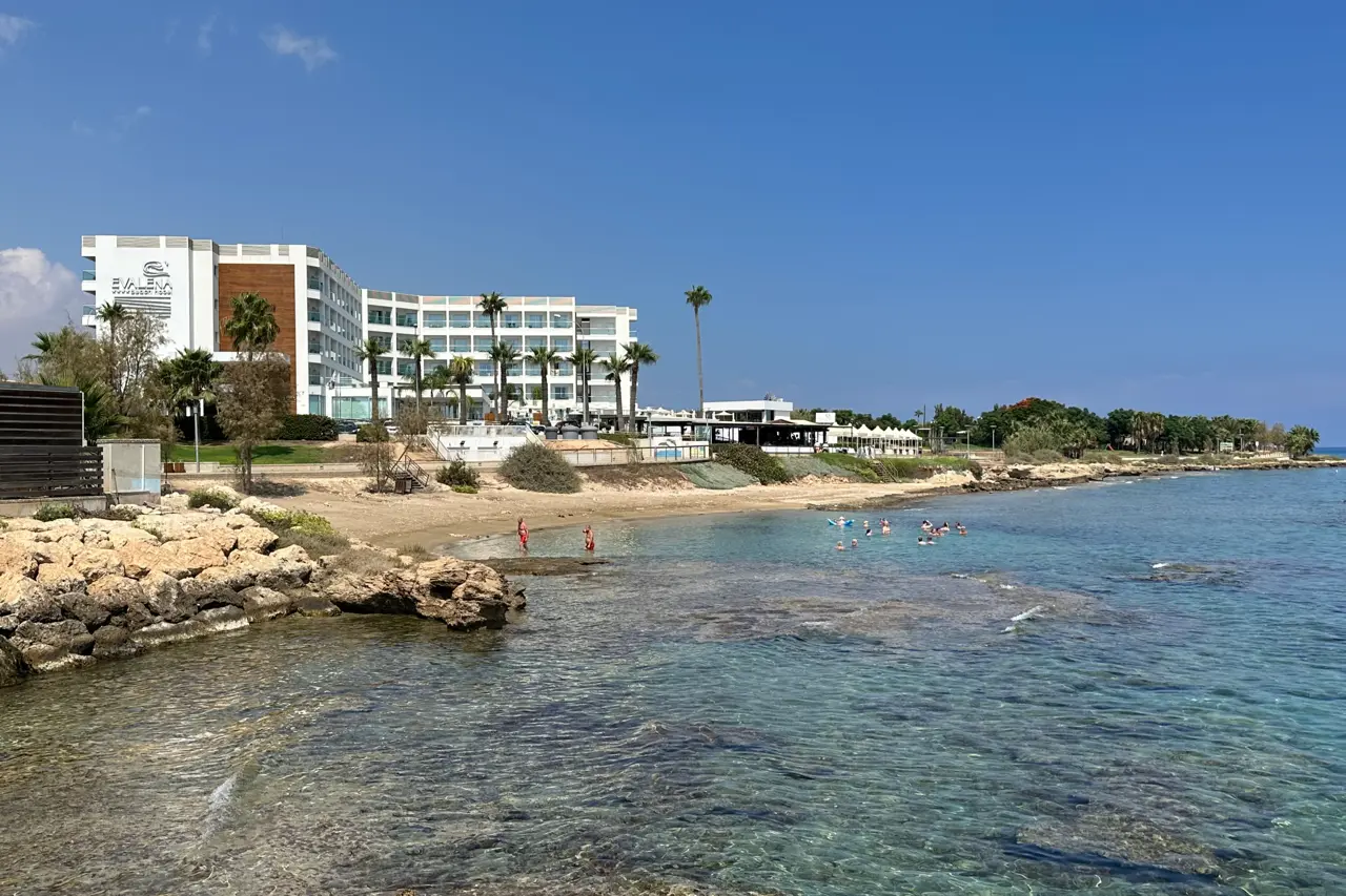 Rocky shore with clear shallow water, a few swimmers by a small sandy beach and a white hotel behind